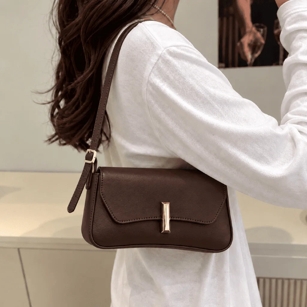 Bolsa Amélia café usada no ombro, com aba curva e detalhe dourado